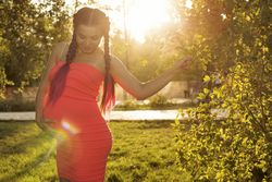 Pregnant female in pink dress standing under trees holding her stomach on the grass on sunny day