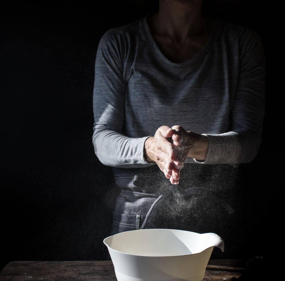 Woman clapping hands in flour near bowl and pack of eggs on table