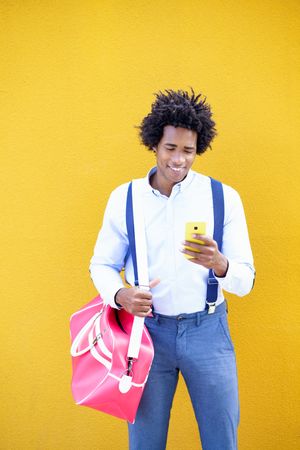 Man wearing suspenders and sports bag smiling at phone in front of yellow wall