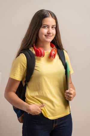 Portrait of a smiling teenage student with a backpack, headphones and a notebook, ready for school.