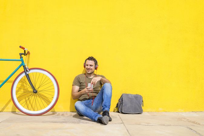 Male sitting in front of yellow wall next to bike with bike and listening to music on smartphone