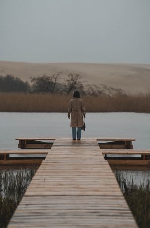 Back view of woman in brown coat standing on wooden dock by the lake