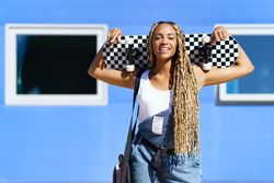 Smiling female in denim overalls standing with skateboard behind head in front of blue building