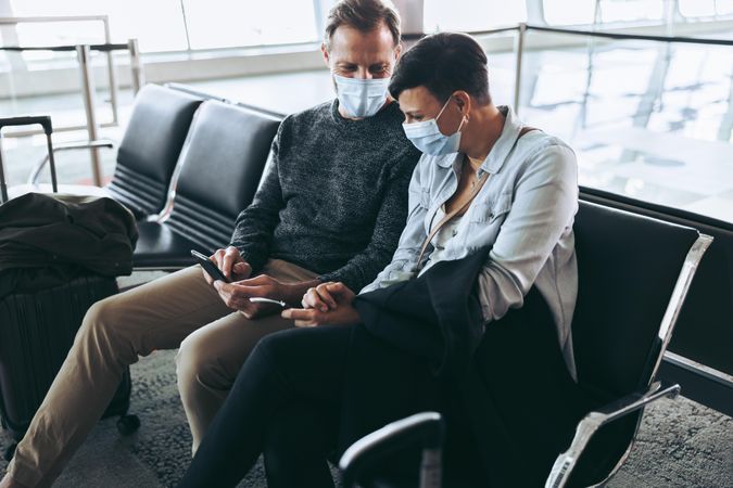 Couple with face masks sitting at airport and using mobile phone to check flight information