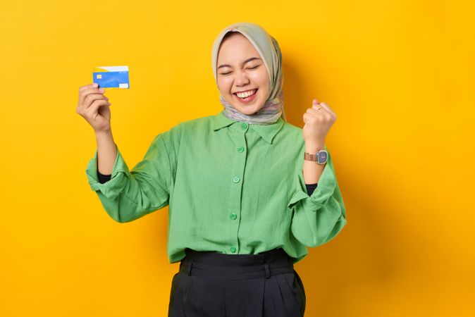 Muslim woman in headscarf and green blouse holding credit card and clenching fist with eyes closed