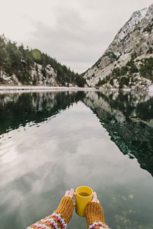 Person with cup near wonderful lake between hills in snow and cloudy sky