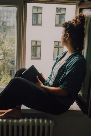 Interested woman reading book at window sill at home