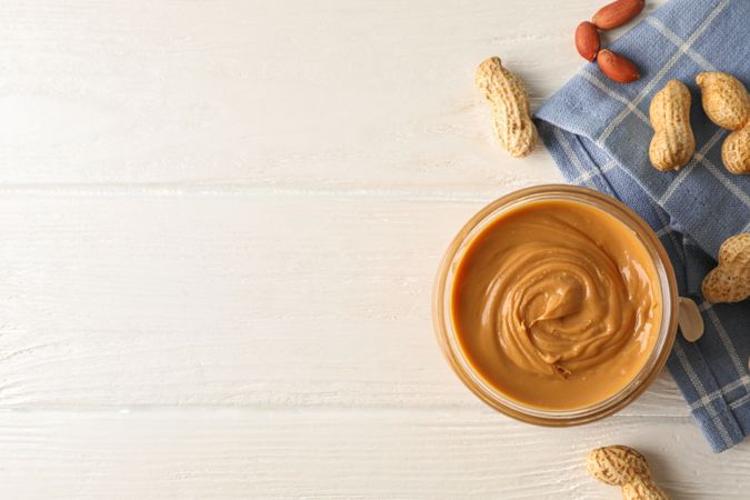 Glass jar with peanut butter, peanut and kitchen towel on wooden background, top view and space for text