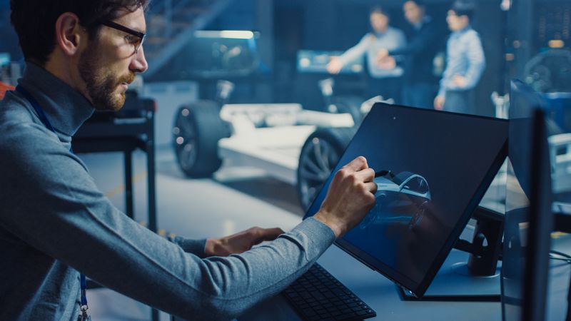 Professional Automotive Graphic Designer is Working on Concept Car Render with a Stylus Pen in a High Tech Innovative Laboratory with a Prototype Car Chassis.