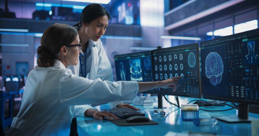 Medical Hospital Research Laboratory: White And Asian Female Scientists Talking And Using Desktop Computer With MRI Scans Of Human Brain.