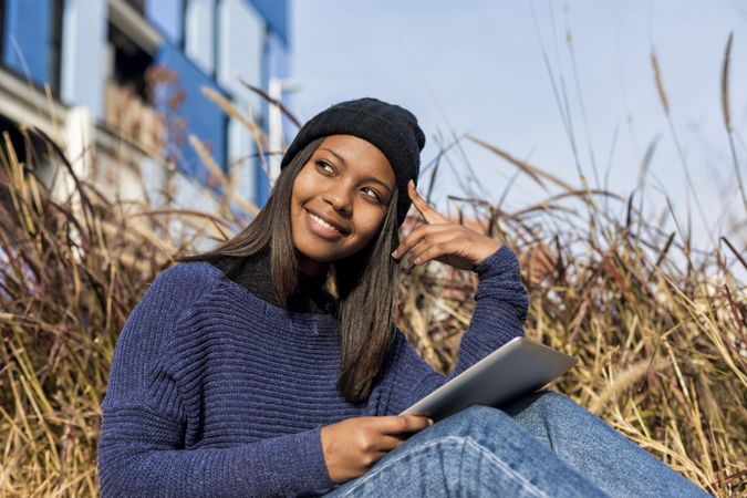 Smiling female in hat and sweater sitting outside with tablet with space for text