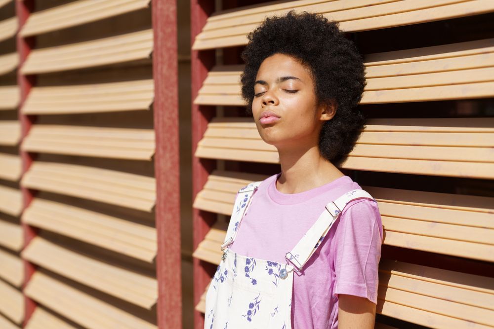Female in overalls leaning on wooden shutters facing the sun with eyes closed