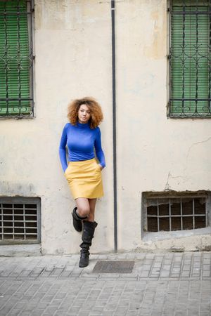 Nonchalant female with curly hair wearing bright blue shirt leaning on wall on street