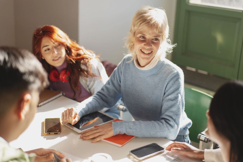 A group of cheerful college students collaboratively using tablets and smartphones for their study session in a bright classroom, showcasing interactive learning