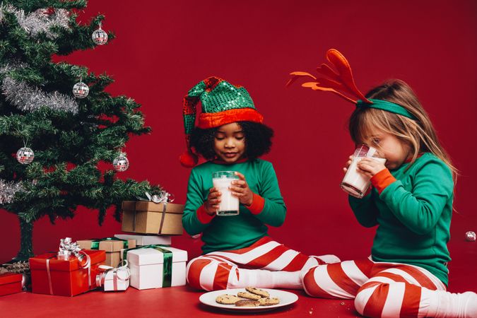 Festive little kids drinking milk sitting beside Christmas tree