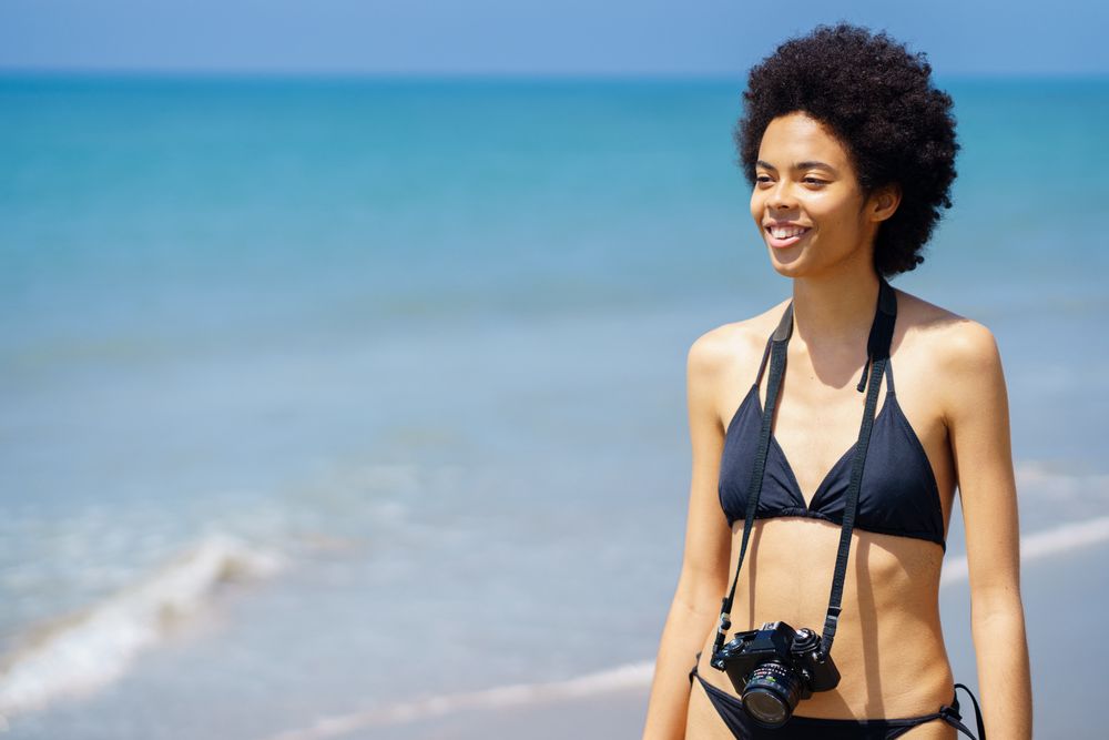 Smiling woman walking along the shore in bikini with camera around her neck