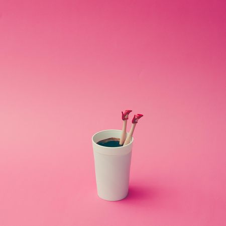 Coffee cup with female doll legs on pink background, with copy space