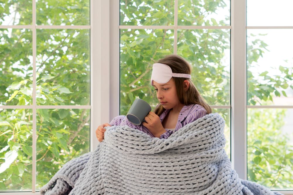 Teenage girl wrapped in grey knitted blanket by window.