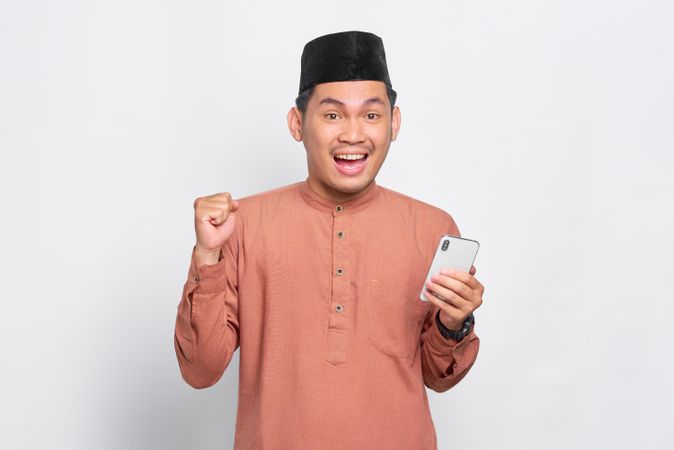 Muslim man in kufi hat holding mobile phone and celebrating something