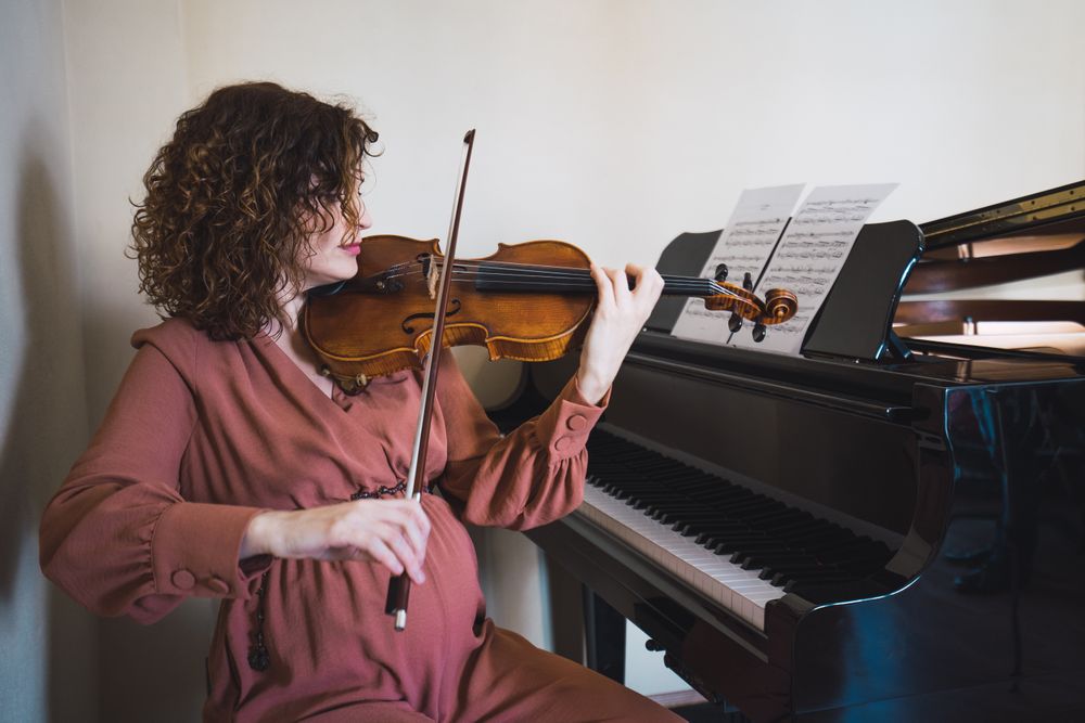 Pregnant woman playing on violin near piano in room