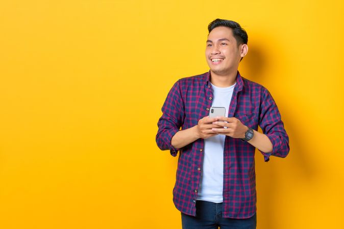 Smiling Asian in plaid shirt  holding smartphone while looking away
