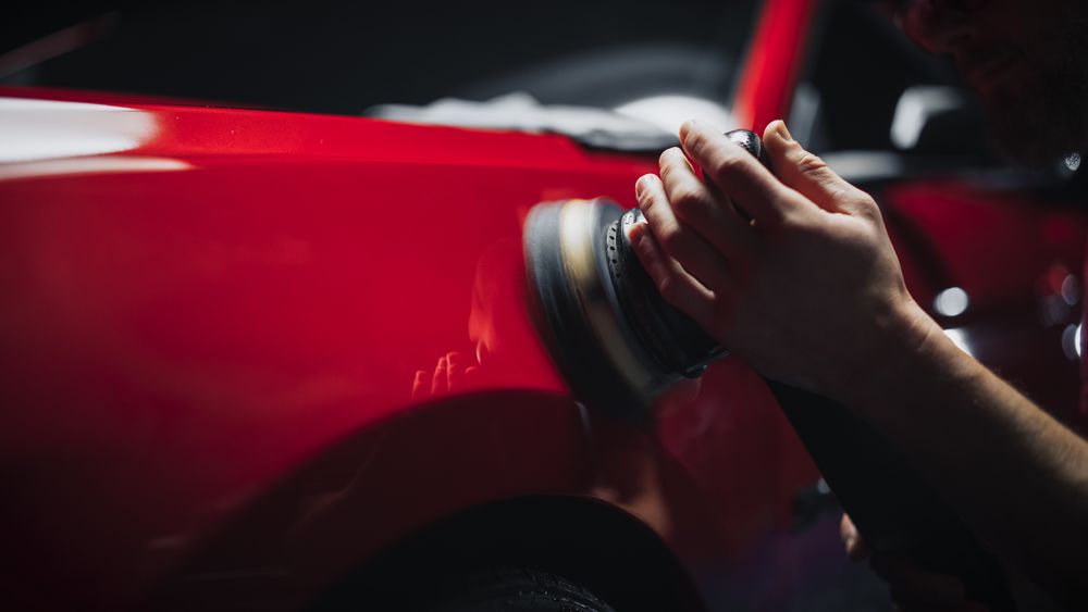 Car Ad Style Photo of a Professional Car Detailer Using an Electric-Powered Polishing Machine to Work on a Fender of a Beautiful Red Sportscar After Washing and Detailing the Vehicle