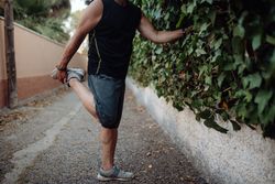 Older man stretching leg holding botanical wall in street