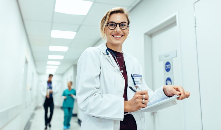Portrait of happy young female physician standing in corridor with clipboard