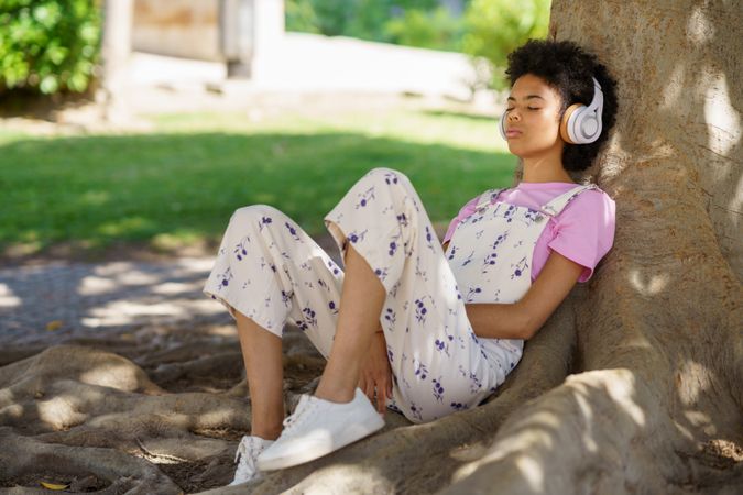 Calm woman in floral coveralls relaxing on roots of large tree in park listening to music