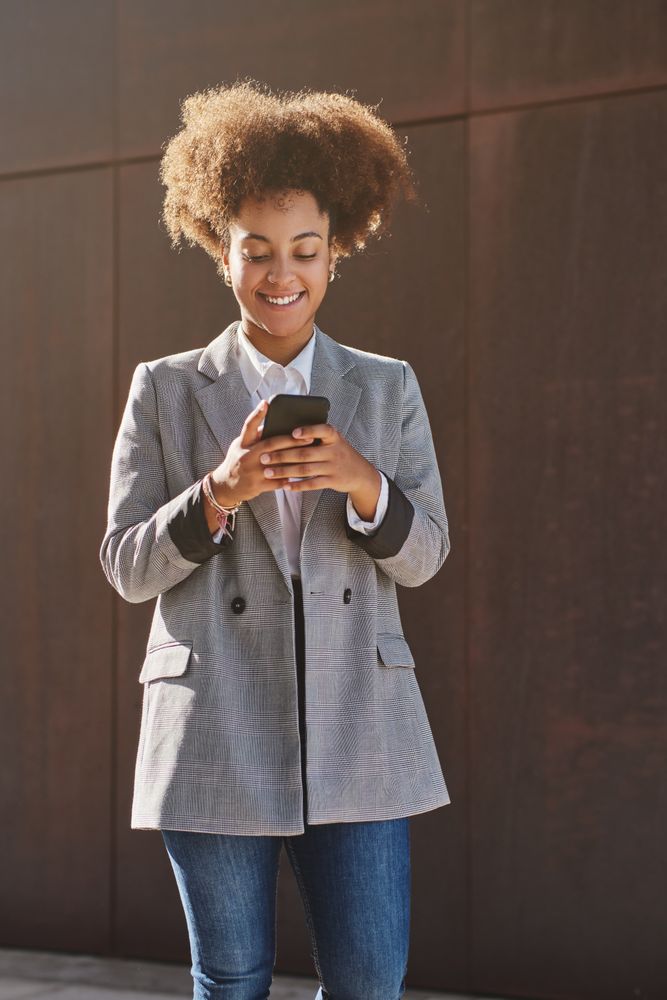 Positive Black female texting on phone near street wall