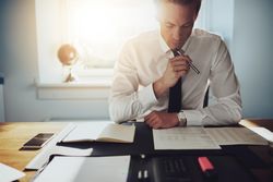 Man in shirt and tie sitting at desk reading over documents in a sunny office