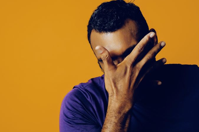 Tired Black man in yellow studio covering his face with his hands