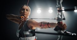 Portrait of a Professional Female Archer Aiming an Arrow at a Target with a Compound Bow. Strong Athlete is Fully Equipped and Protected with Chest and Arm Guards, Preparing For Tournament