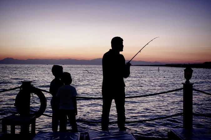 Silhouette of man holding fishing rod and two boys standing by seashore during sunset