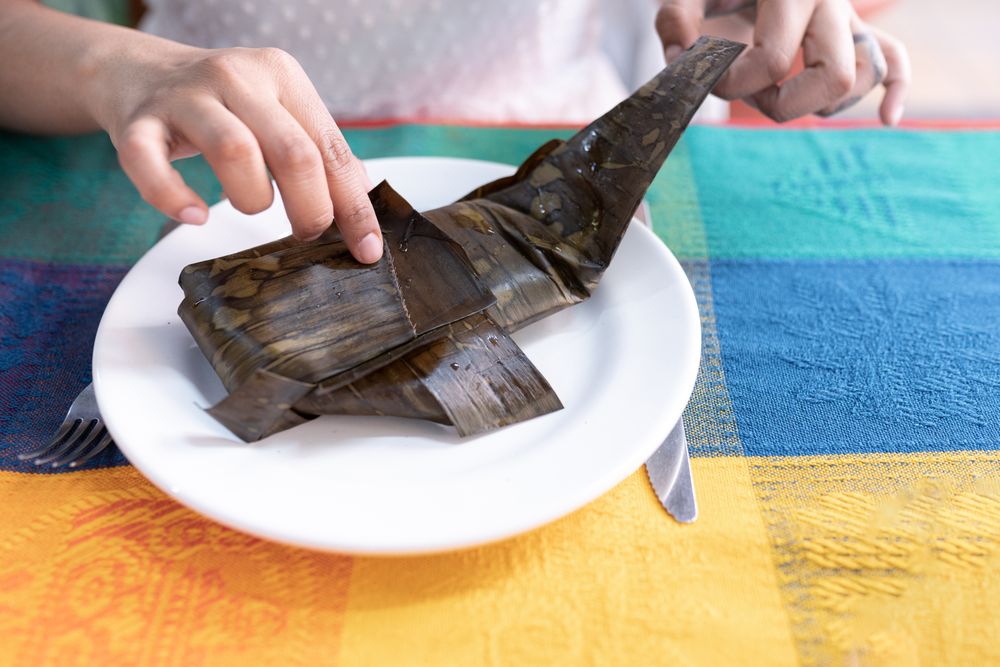 A young Hispanic woman is opening a meat tamale wrapped with a plant leaf in a restaurant