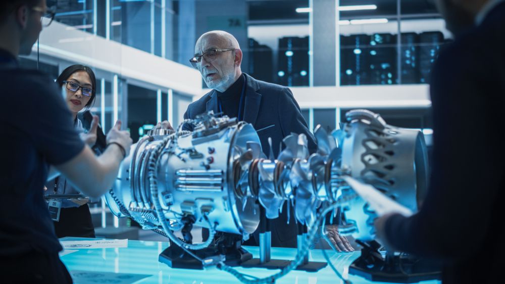 Diverse Multiethnic Team of Industrial Engineers Gathered Around a Table With a Prototype Turbofan Engine.