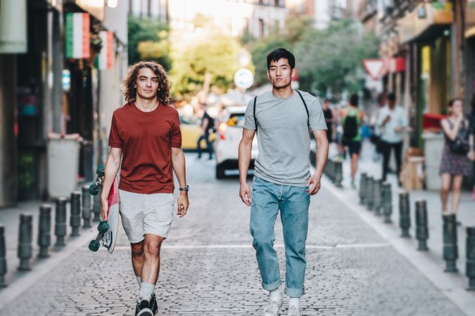 Cool multiracial male friends chatting while walking on street with longboard