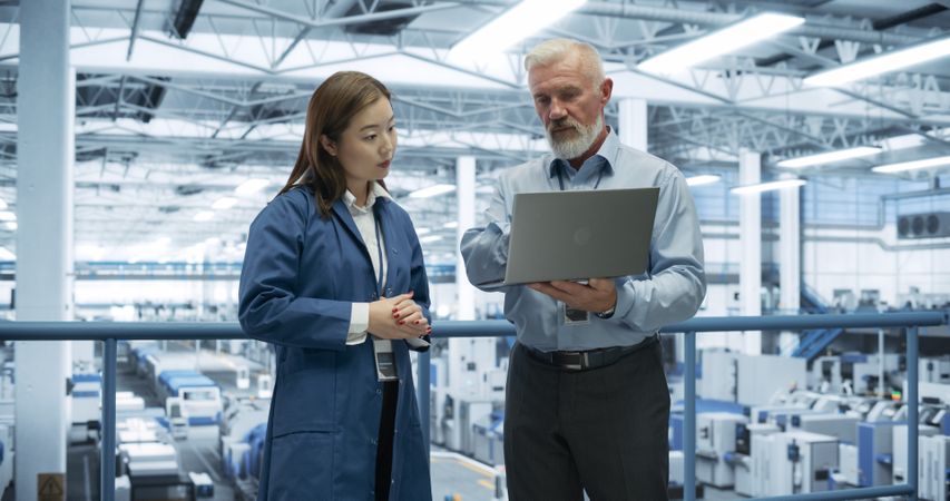 Experienced Male and Female Engineers Standing on a Platform, Using Laptop Computer and Discussing Production at a Modern Automated Electronics Manufacture with AI Support