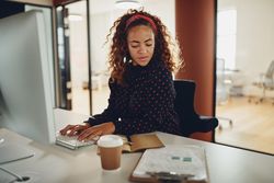 Woman with curly hair checking her notes and a coffee at computer at work