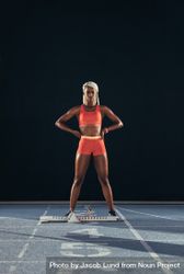 Female athlete standing beside a starting block on running track on a dark background 5XpYM5