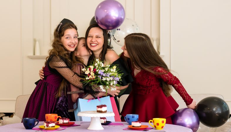 Woman hugging girls at birthday party with cake and flowers.