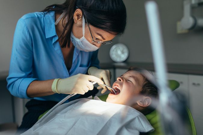Female dentist examining teeth of little boy in dental clinic using dental tools