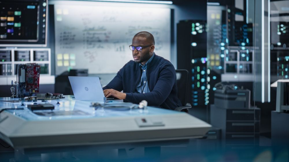 Portrait of a Male Specialist Using Laptop Computer to Improve Electronic Circuit Board in Modern Office. Black Engineer Thinking and Typing Ideas for Artificial Intelligence Project