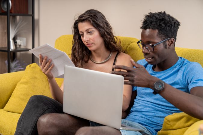A diverse couple at home reviewing documents and using a laptop to manage their household finances together.