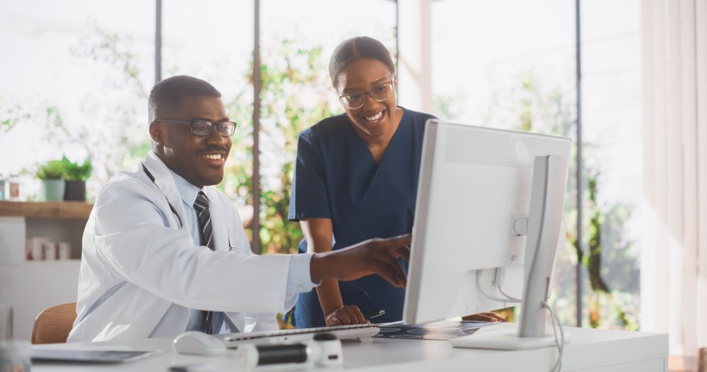 Black Nurse and Doctor Having a Casual Conversation, Using Desktop Computer, Laughing and Smiling During a Break at Work