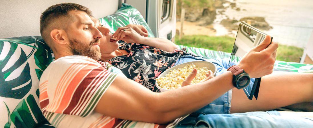 Loving male and female reclining on bed in motorhome with popcorn and digital tablet