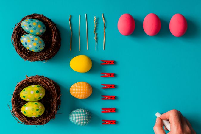 Bright Easter eggs, birds nests and clothes pins on cyan paper with hand holding pen