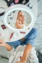 Cheerful woman smearing cream on face with brush opposite mirror