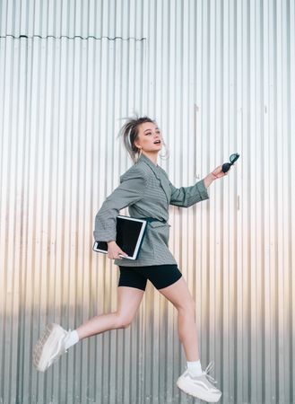 Woman with tablet in stylish suit jumping nearby wall holding sunglasses