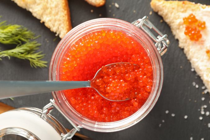Composition with caviar in glass jar, bread, spoon and cutting board, top view